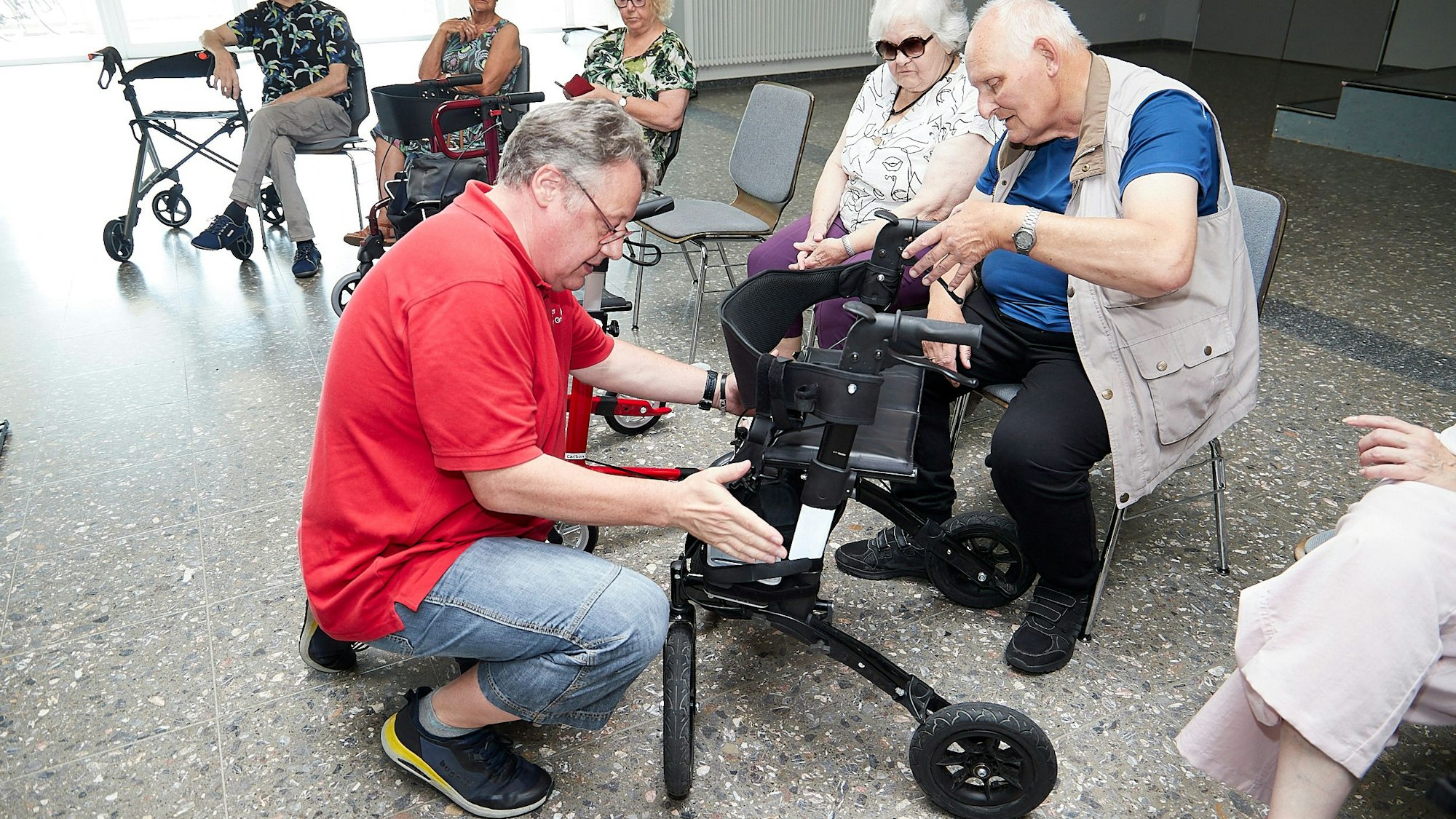 Ein Mann kniet vor einem Rollator und stellt die Bremse ein. Der Rollatorbesitzer sitzt auf einem Stuhl und schaut zu.