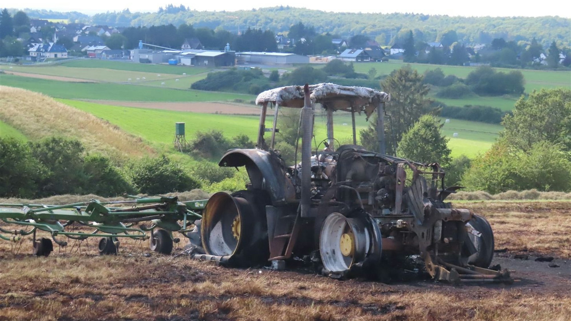Ein Traktor steht völlig ausgebrannt auf einer Wiese, ein Heuwender ist angeschirrt.
