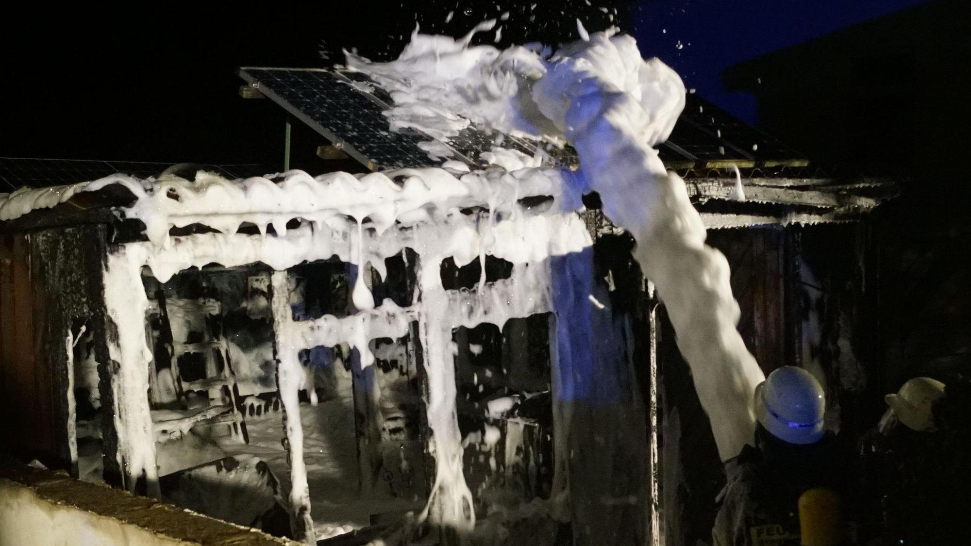 Feuerwehrleute spritzen Schaum auf die Reste der ausgebrannten Gartenlaube.
