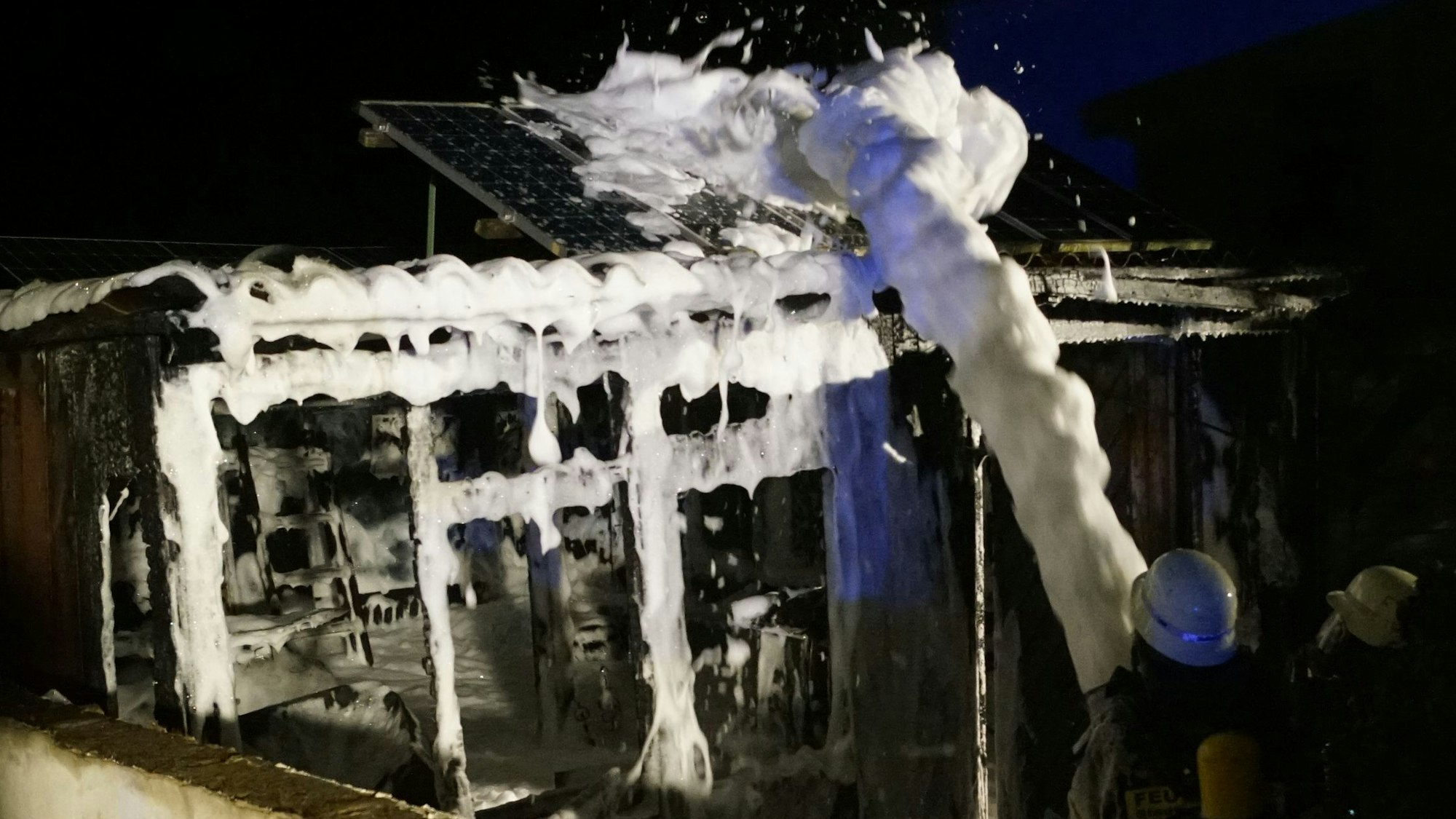 Feuerwehrleute spritzen Schaum auf die Reste der ausgebrannten Gartenlaube.