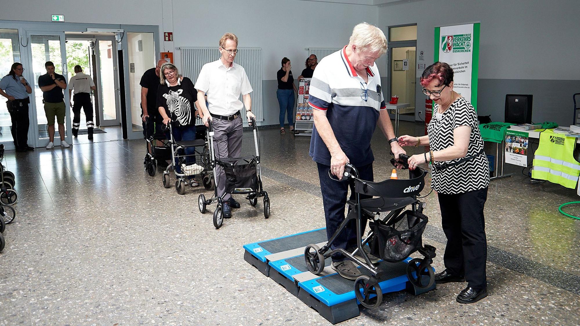 Ein Mann probiert aus, wie es ist, mit einem Rollator über ein Podest zu fahren. Die Trainerin gibt ihm Tipps.