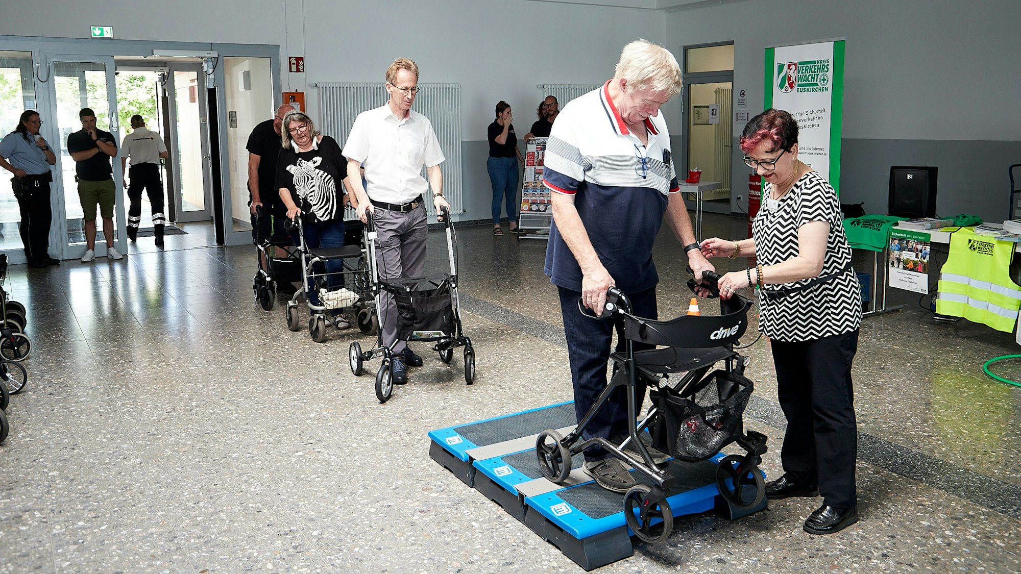 Ein Mann probiert aus, wie es ist, mit einem Rollator über ein Podest zu fahren. Die Trainerin gibt ihm Tipps.
