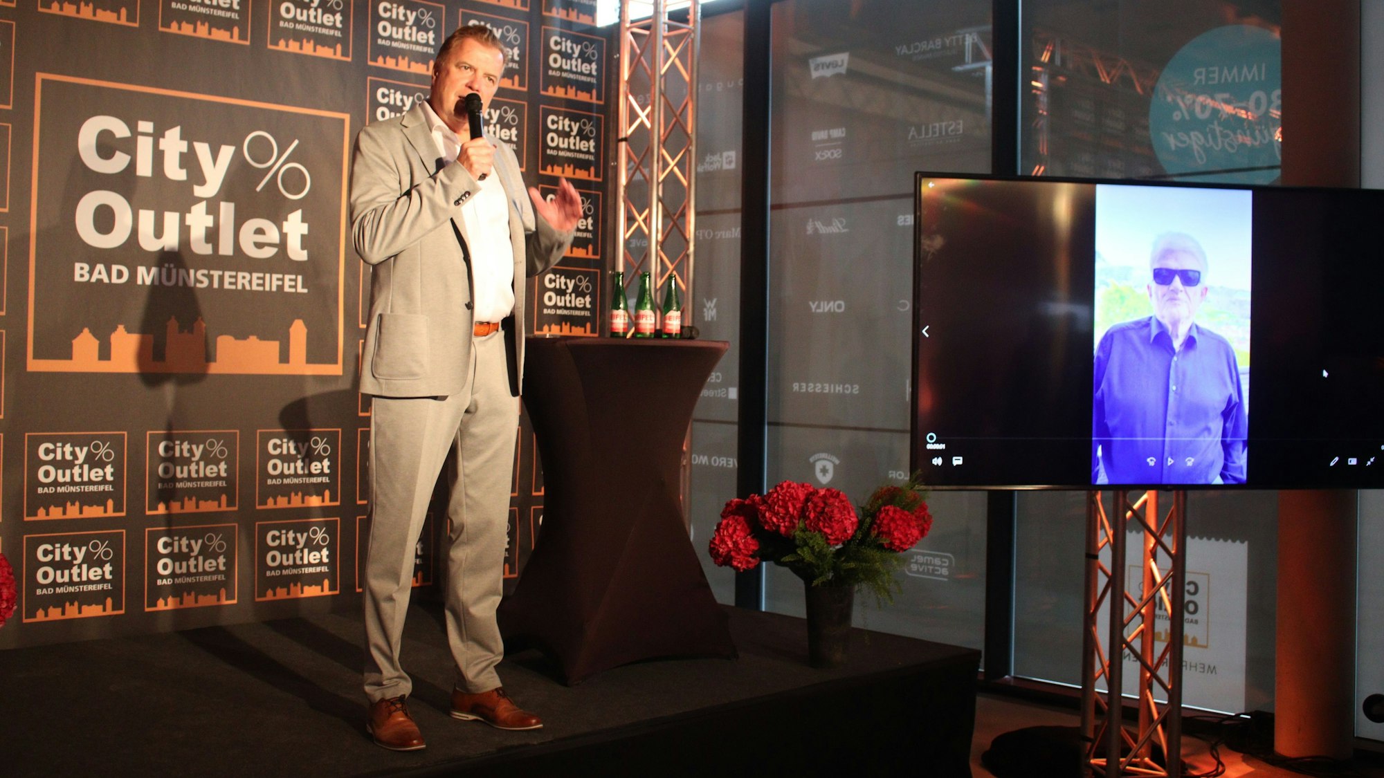 Marc Brucherseifer steht auf der Bühne und spricht in ein Mikrofon. Auf einem Bildschirm ist Heino zu erkennen.