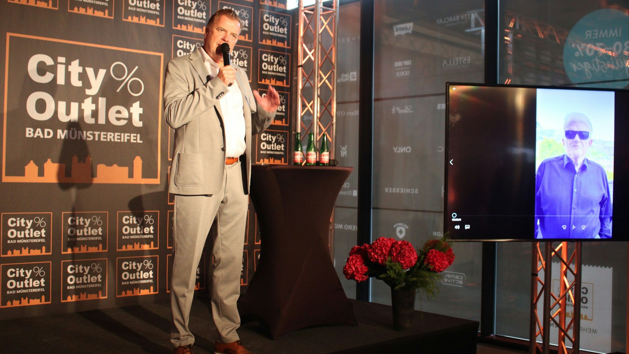 Marc Brucherseifer steht mit Mikrofon auf der Bühne zur Eröffnung des neuen Gebäudes. Auf einem Bildschirm ist Heino zu sehen.