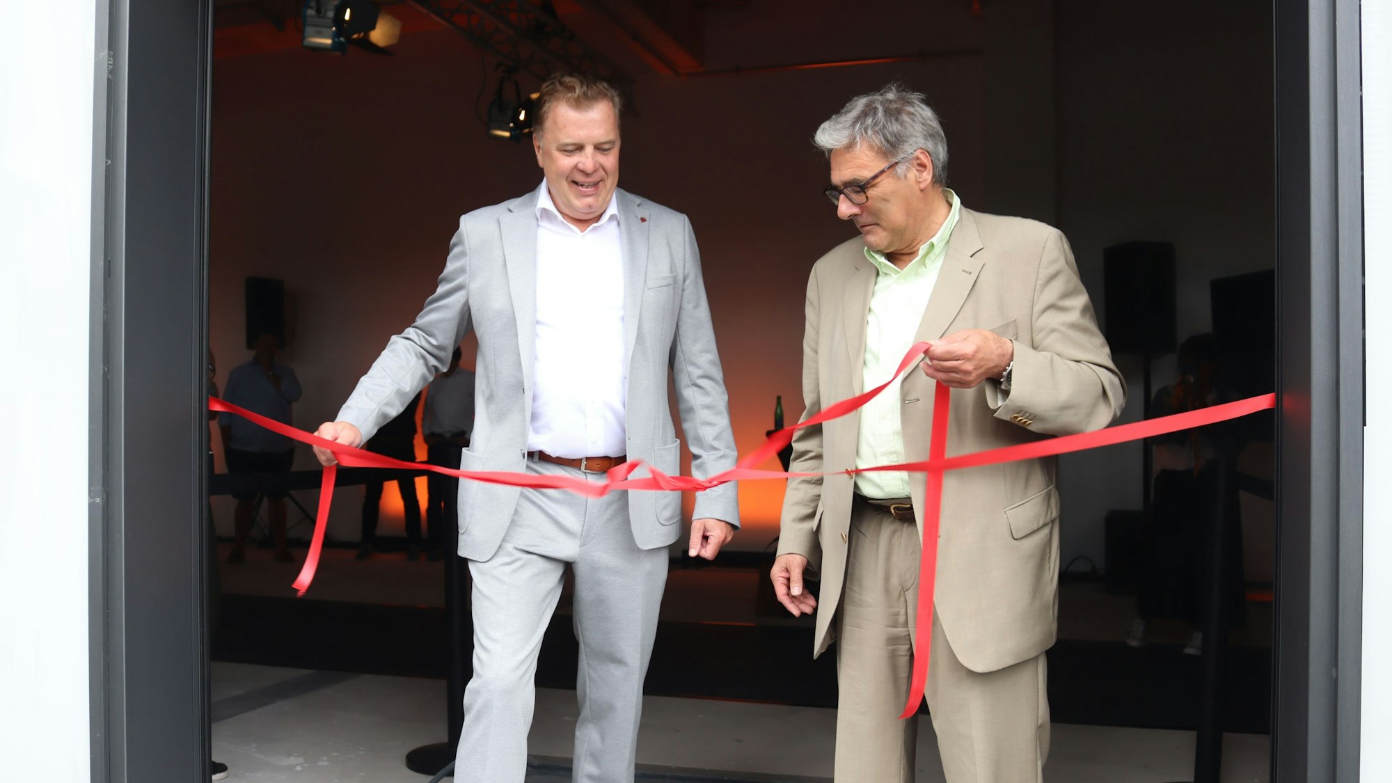 Marc Brucherseifer und Ludger Müller stehen im Eingang eines Geschäfts und ziehen an den Enden einer roten Schleife.