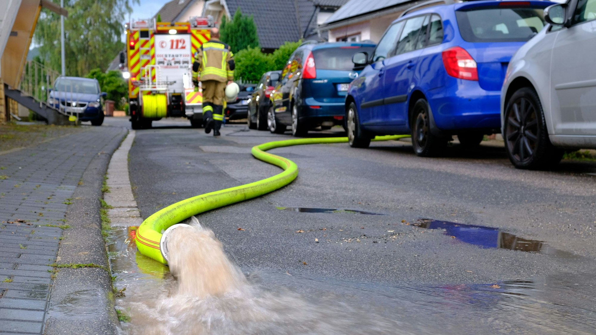 Wasser sprudelt aus einem gelben Feuerwehrschlauch: Helfer der Freiwilligen Feuerwehr pumpen nach einem Starkregenereignis am 13.8.24 Keller in der Rehgasse in Kommern aus.