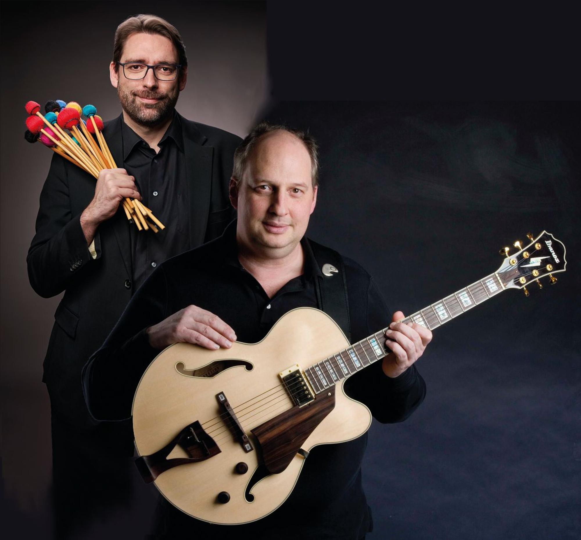Zwei Musiker, einer hat Schlägel in der Hand, der andere hält Gitarre fest.