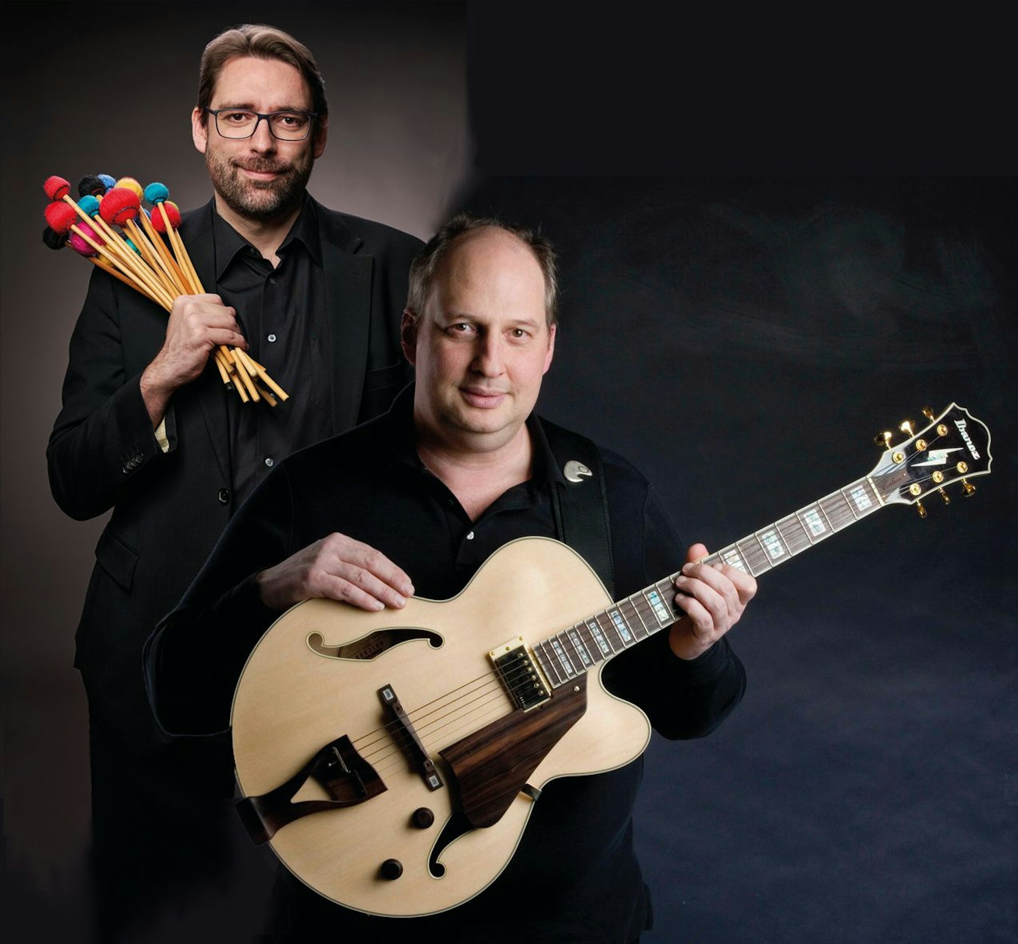 Zwei Musiker, einer hat Schlägel in der Hand, der andere hält Gitarre fest.
