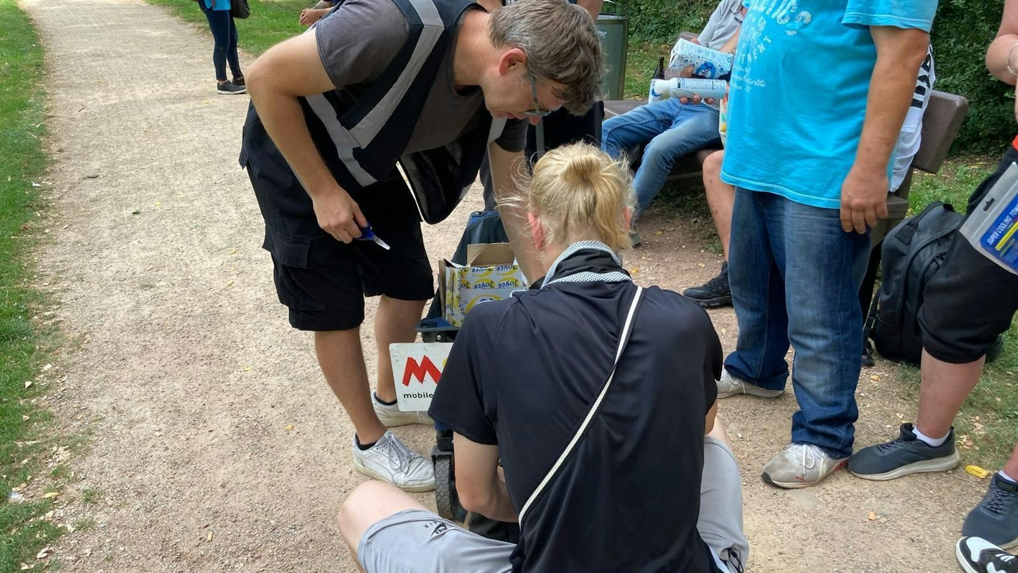 Ein Mann hockt vor einem Bollerwagen auf einem Weg in einem Park, während ein anderer Mann in den Wagen greift.