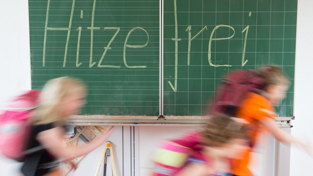 Eine Schultafel, auf der hitzefrei steht. Schülerinnen und Schüler rennen vor der Tafel verschwommen von links nach rechts.