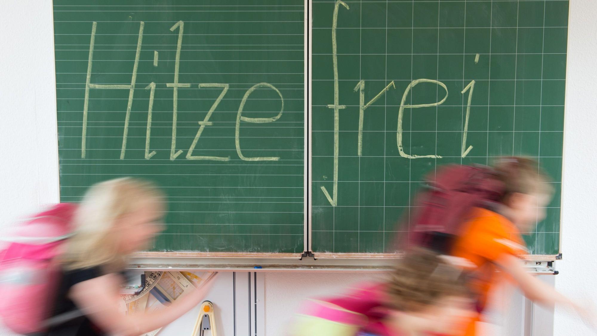 Eine Schultafel, auf der hitzefrei steht. Schülerinnen und Schüler rennen vor der Tafel verschwommen von links nach rechts.