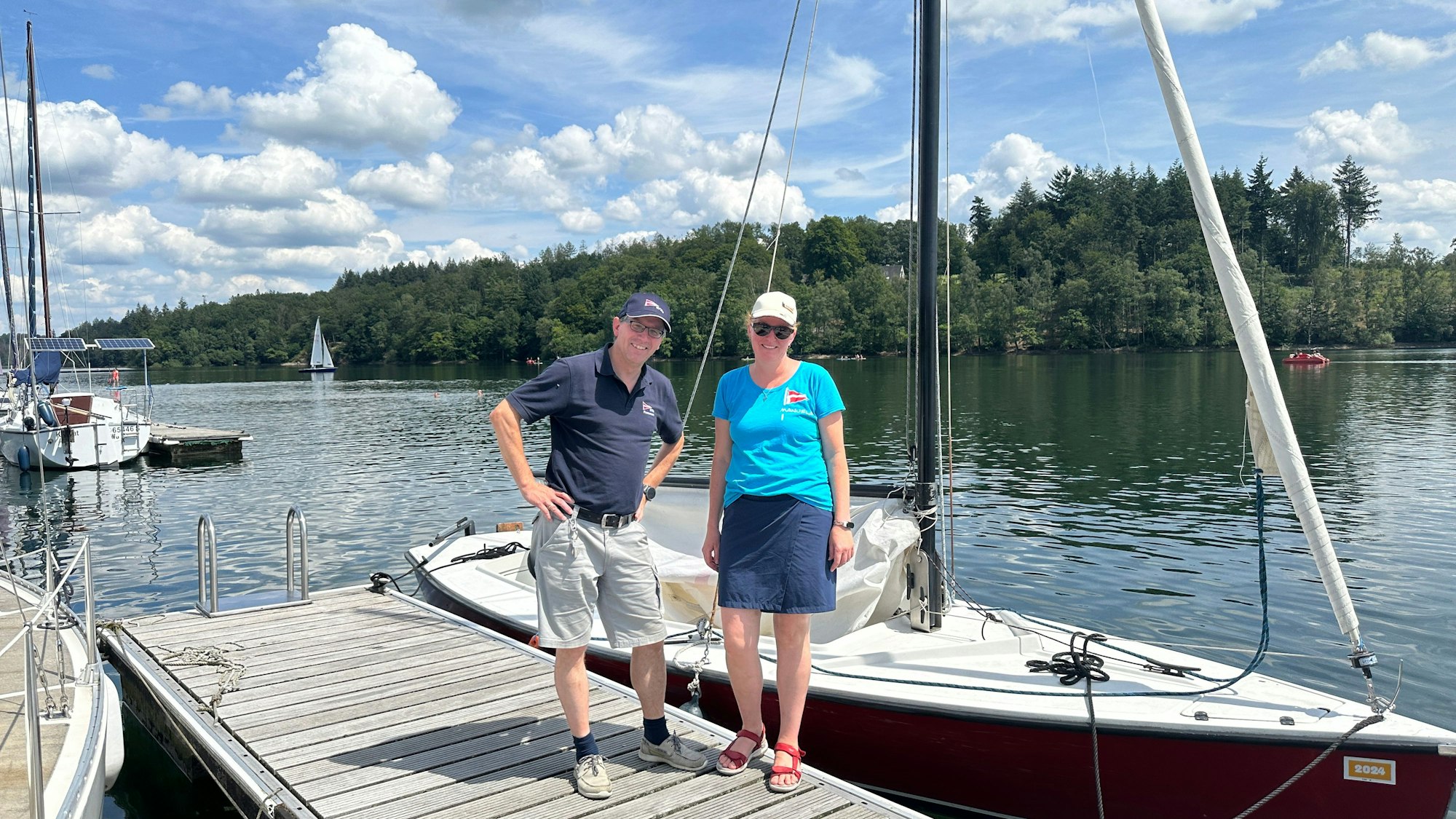 Das Foto zeigt zwei Personen, die auf einem Steg stehen, dahinter ein Segelboot