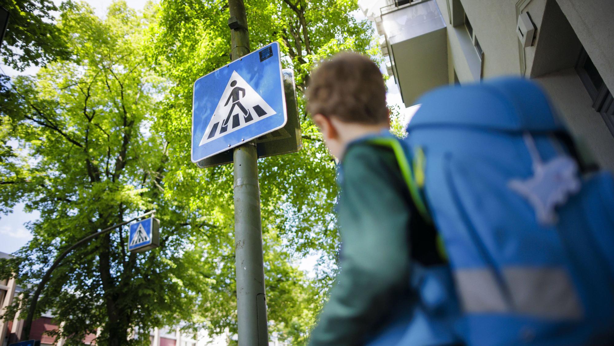 Symbolfoto zum Thema Schulkind im Straßenverkehr. Ein sechsjähriger Junge steht mit seinem Schulranzen auf dem Rücken vor dem Schild eines Fußgängerüberweges.