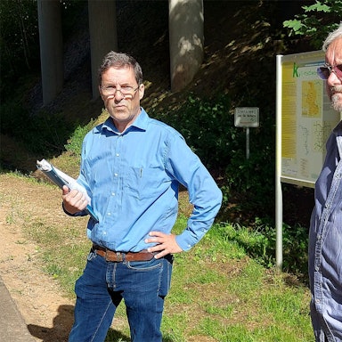 Zwei Männer in blauen Hemden stehen an einem bewaldeten Weg.