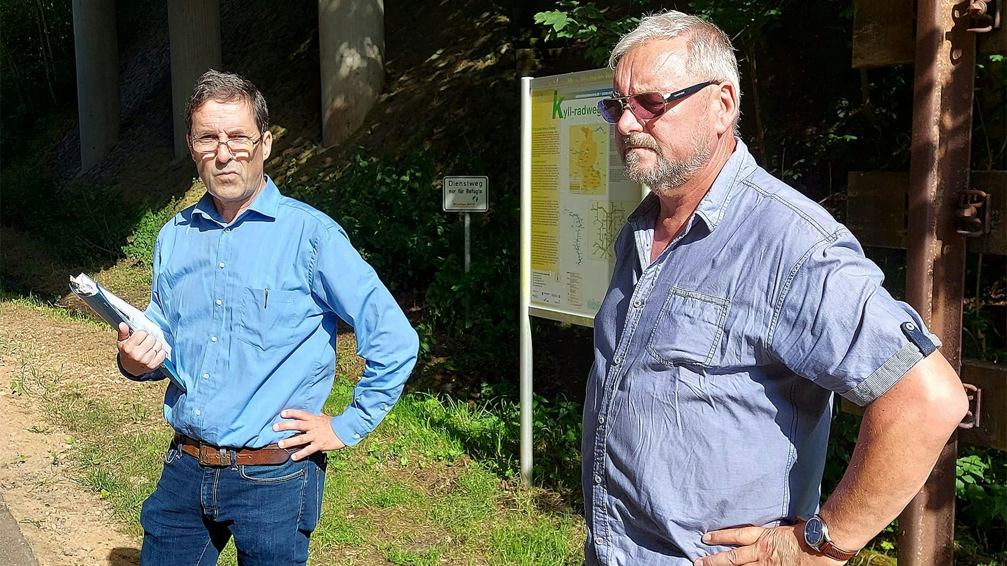 Zwei Männer in blauen Hemden stehen an einem bewaldeten Weg.