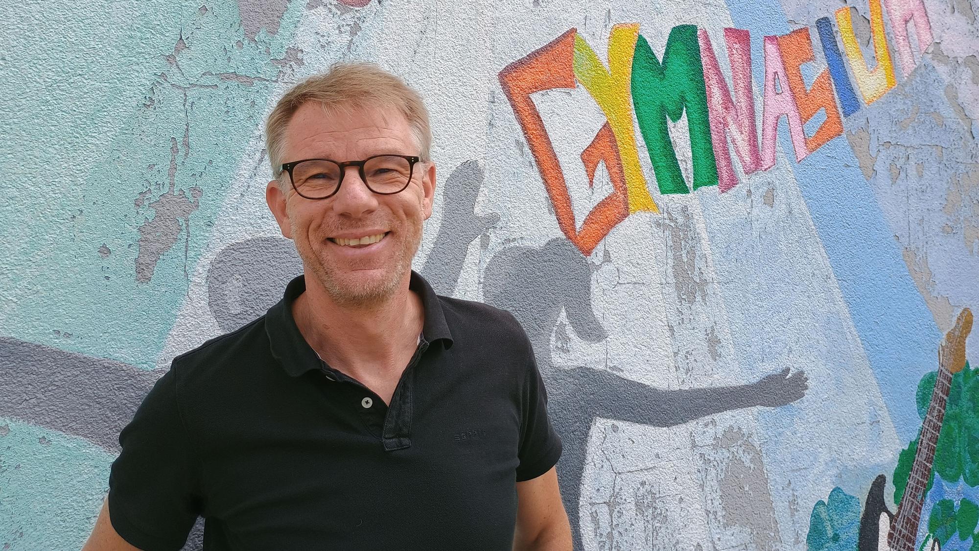 Oliver Großmann trägt ein schwarzes Polo-Hemd. Hinter ihm ist eine Wand bemalt, etwa mit einer eine bunte Aufschrift mit dem Wort Gymnasium.