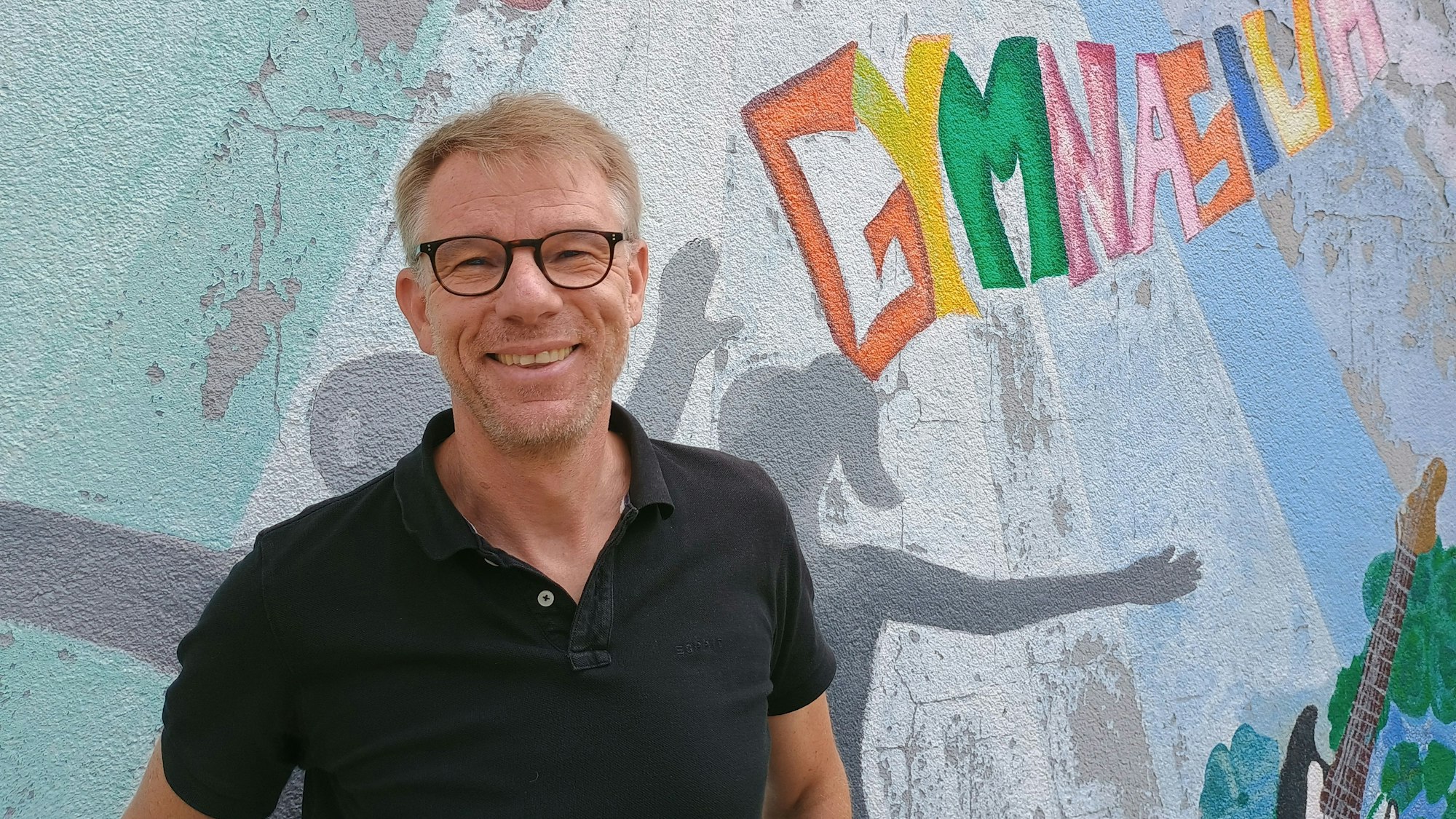 Oliver Großmann trägt ein schwarzes Polo-Hemd. Hinter ihm ist eine Wand bemalt, etwa mit einer eine bunte Aufschrift mit dem Wort Gymnasium.