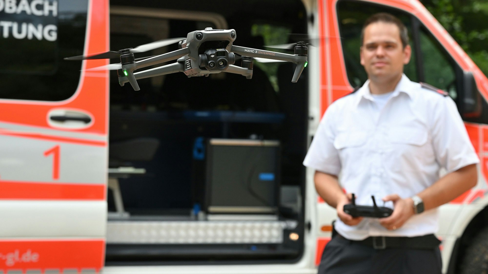 Marco Braun hält das Steuergerät der ersten Feuerwehrdrohne im Kreis und steuert damit das Fluggerät im Vordergrund. Im Hintergrund ist der neue Einsatzleitwagen zu sehen.