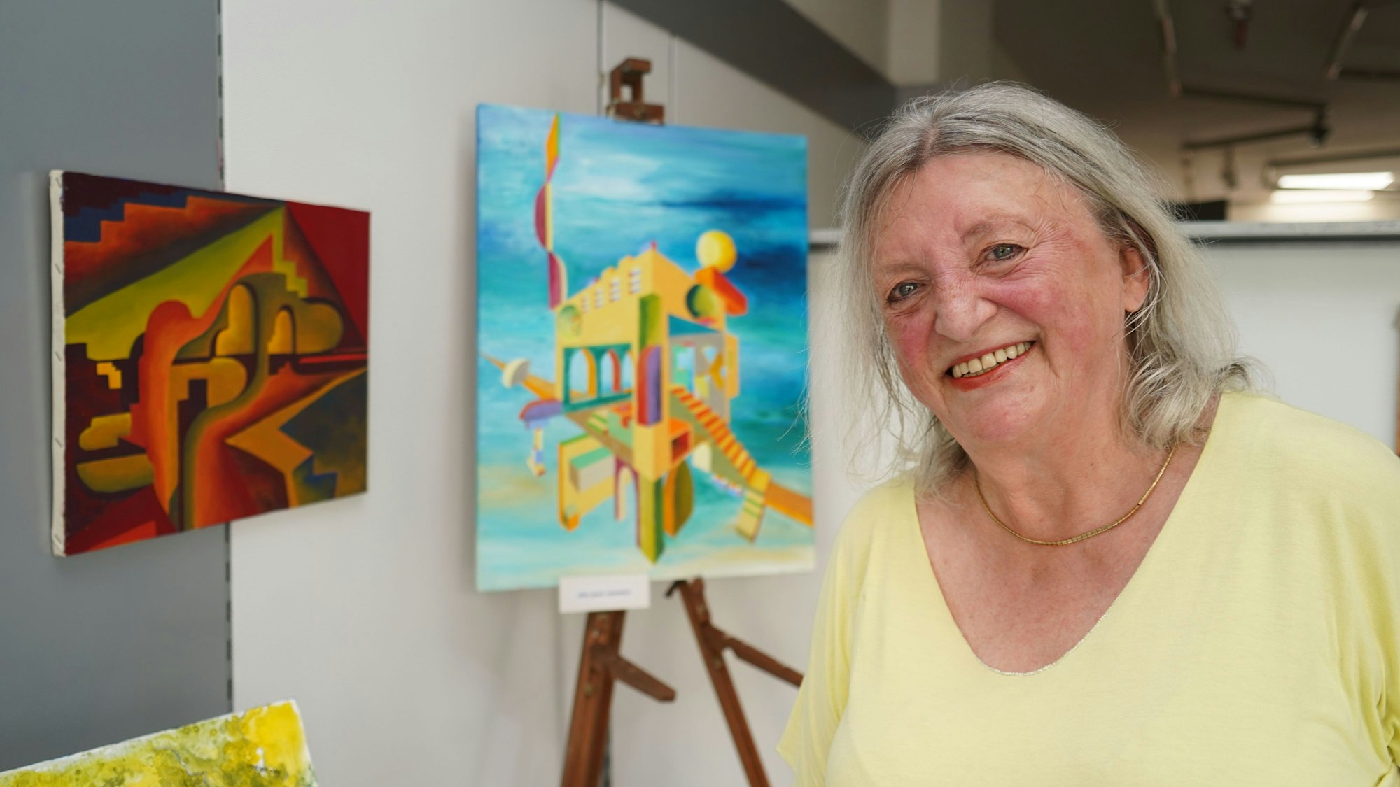 Eine Frau lächelt in die Kamera, hinter ihr steht eine Staffelei mit einem ihrer Bilder, ein anderes hängt an der Wand.