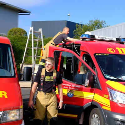 Zwei Feuerwehrleute stehen an einem Mannschaftstransportwagen, auf der Beifahrertür ist das Wappen der Stadt Mechernich zu sehen. Sie bereiten sich darauf vor, die Bevölkerung mit Lautsprecherdurchsagen zu informieren.