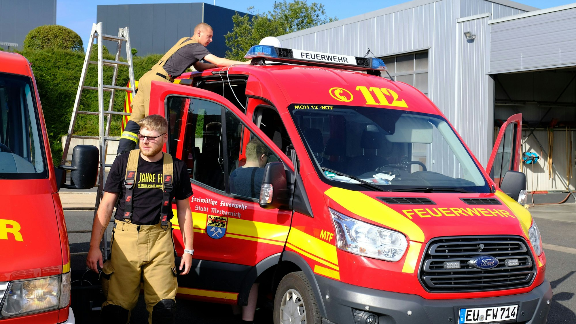 Zwei Feuerwehrleute stehen an einem Mannschaftstransportwagen, auf der Beifahrertür ist das Wappen der Stadt Mechernich zu sehen. Sie bereiten sich darauf vor, die Bevölkerung mit Lautsprecherdurchsagen zu informieren.