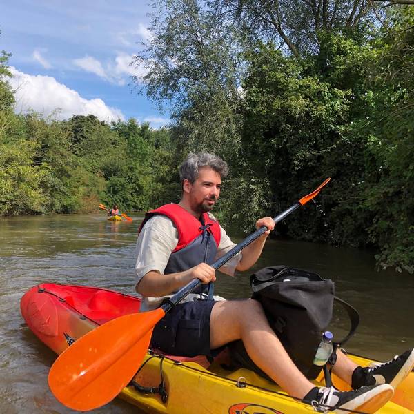 Auf dem Bild ist ein wasserscheuer Journalist zu sehen, der nach bestem Gewissen auf der Erft paddelt. Das Kajak ist vorne gelb und hinten rot. Im Hintergrund Büsche und Bäume am Ufer des Wassers.