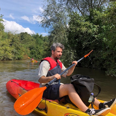 Auf dem Bild ist ein wasserscheuer Journalist zu sehen, der nach bestem Gewissen auf der Erft paddelt. Das Kajak ist vorne gelb und hinten rot. Im Hintergrund Büsche und Bäume am Ufer des Wassers.