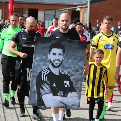 Die Spieler gehen von der Kabine auf den Platz. Einer trägt ein schwarz-weißes Foto des verunglückten Spielers.