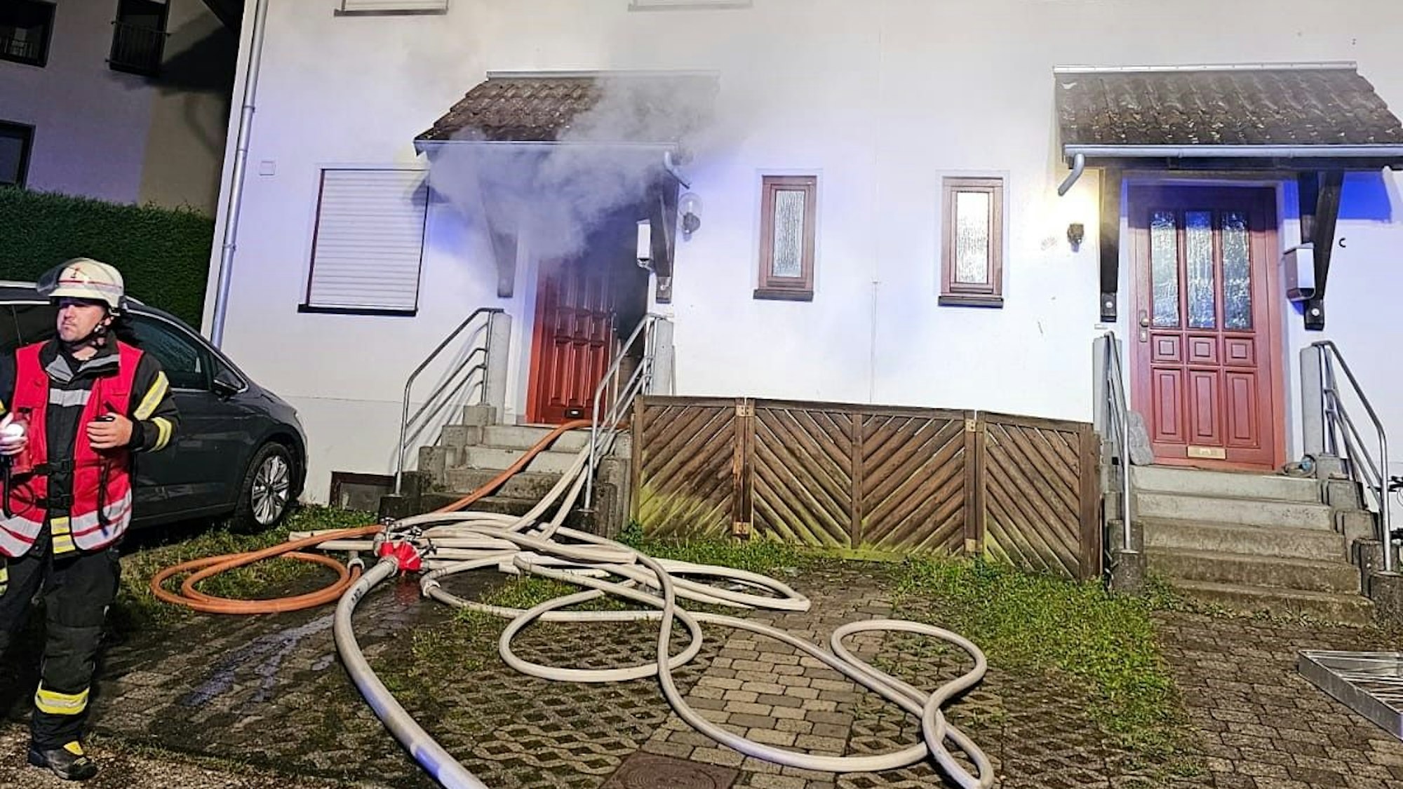 Ein Feuerwehrmann steht vor einem Haus in Bad Honnef-Brüngsberg, Schläuche sind verlegt, dicker Rauch zieht aus der offenen Haustür.