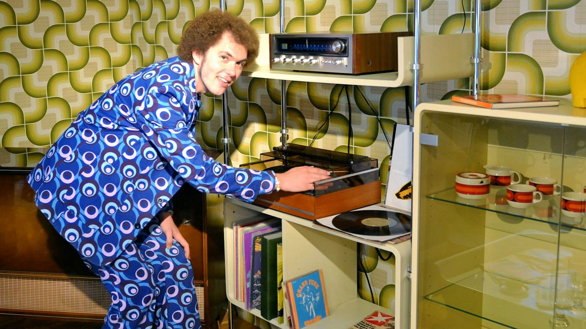 Tobias Schmitz trägt einen bunt gemusterten Anzug und bedient die Musikanlage bei der Ausstellung authentischer Möbelstücke aus den 1970er-Jahren.