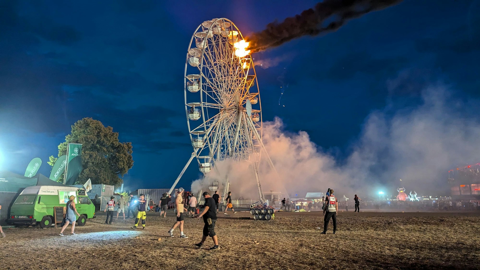 Besucher des Highfield-Festivals und Helfer blicken auf ein Riesenrad, an dem Flammen zu sehen sind. Auf dem Highfield-Festival am Störmthaler See ist ein Feuer an einem Riesenrad ausgebrochen.