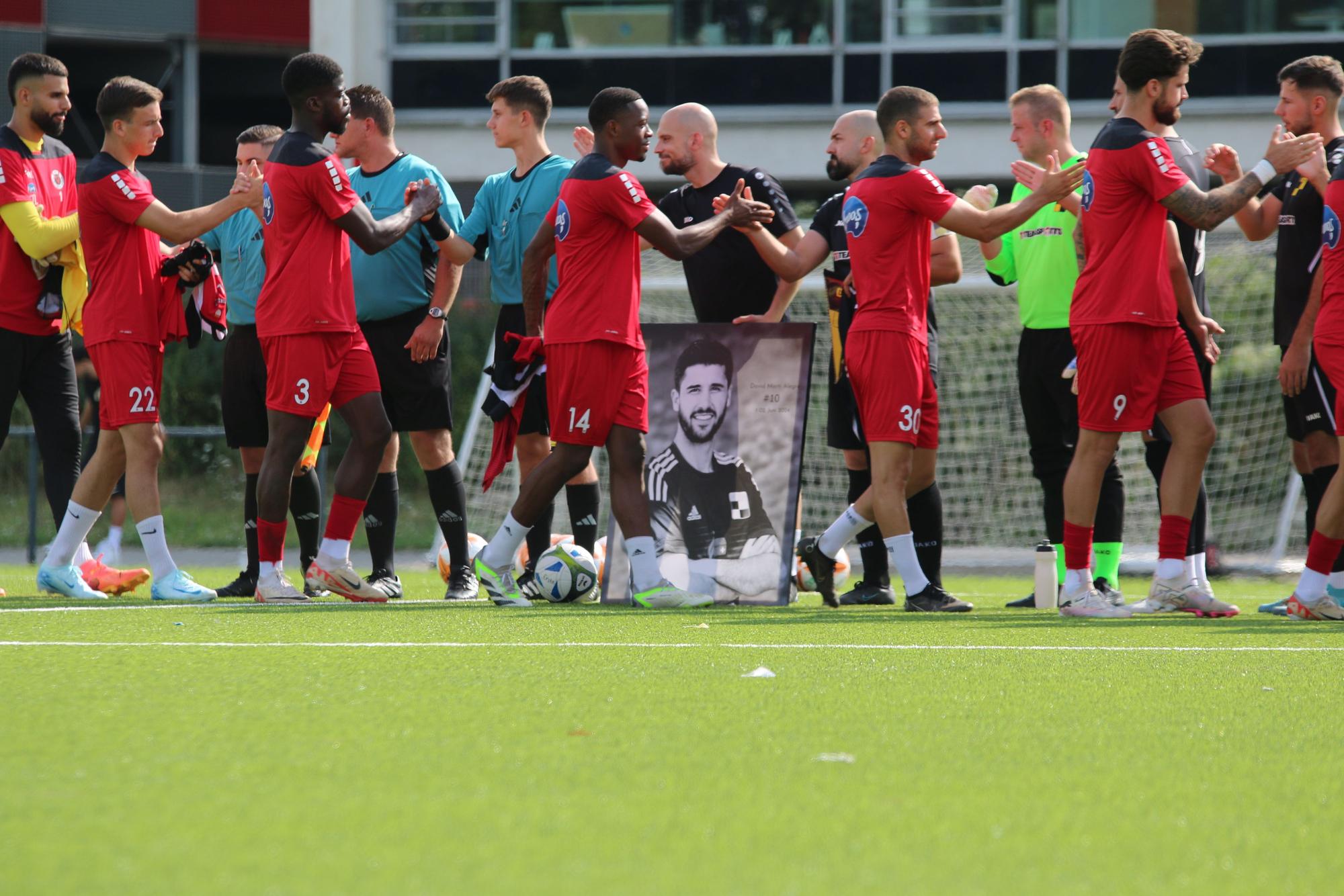 Die Spieler geben sich vor dem Anpfiff die Hand, das Bild von David Marti Alegre steht dabei vor einem der Spieler.