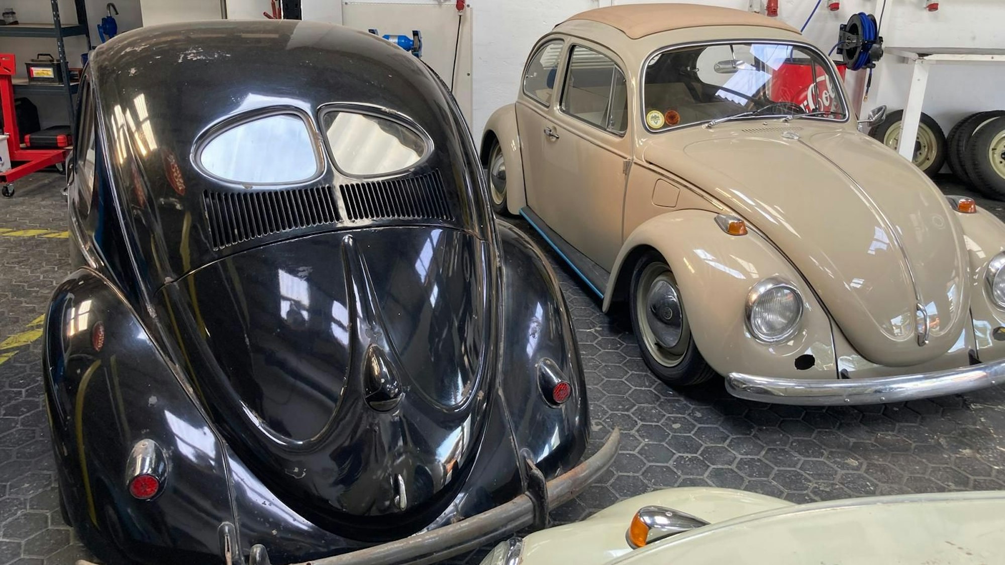 Ein Brezelkäfer steht mit der Rückseite in Richtung Kamera in einer Werkstatt neben einem anderen VW-Oldtimer.
