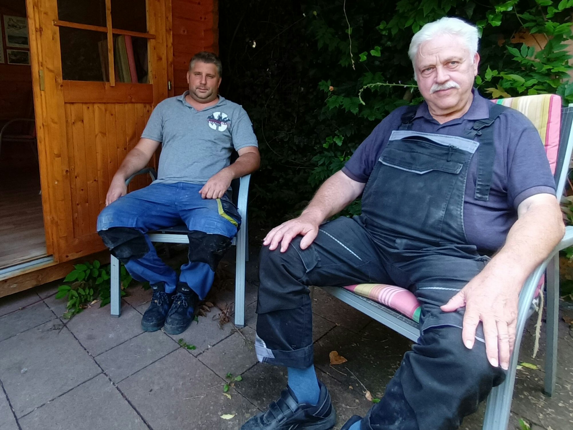 Zwei Männer in Arbeitssachen vor einer Holzhütte sitzend