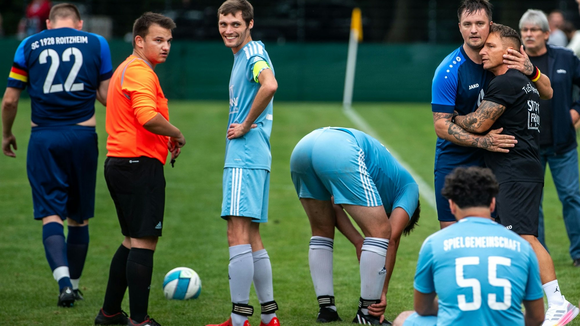 Das Bild zeigt die Szene nach dem Platzverweis. Der Schiedsrichter ist im Gespräch mit einem Spieler, Michal Jedrowski und Michael Kandel umarmen sich.