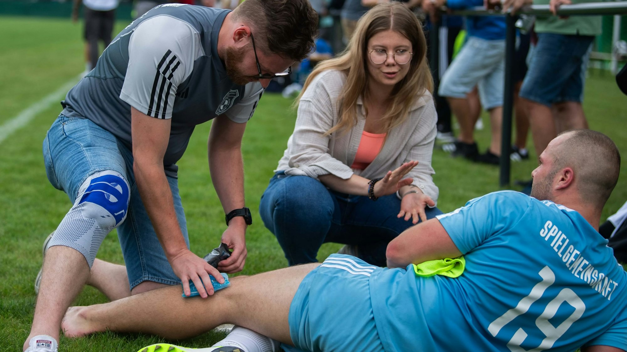 Das Bild zeigt Florian Lanzerath an der Seitenlinie auf der Seite liegend. Von einem Betreuer bekommt er ein Kühlpack aufs Knie gedrückt.