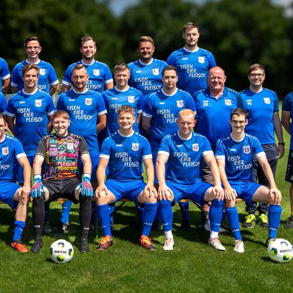 Die Mannschaft des TuS Dom-Esch posiert in blauen Trikots auf einem Rasenplatz. Im Hintergrund stehen unscharf Bäume.