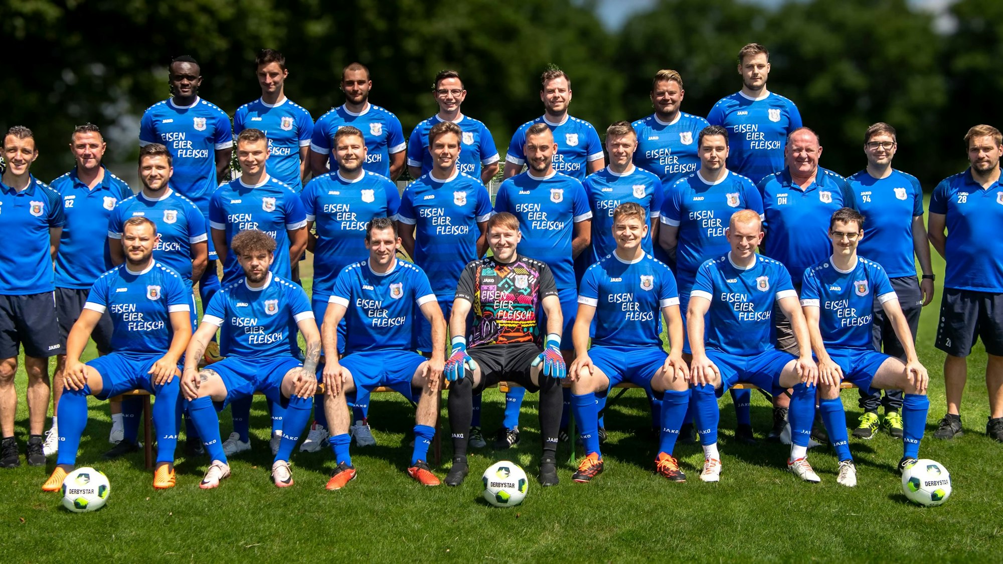 Die Mannschaft des TuS Dom-Esch posiert in blauen Trikots auf einem Rasenplatz. Im Hintergrund stehen unscharf Bäume.