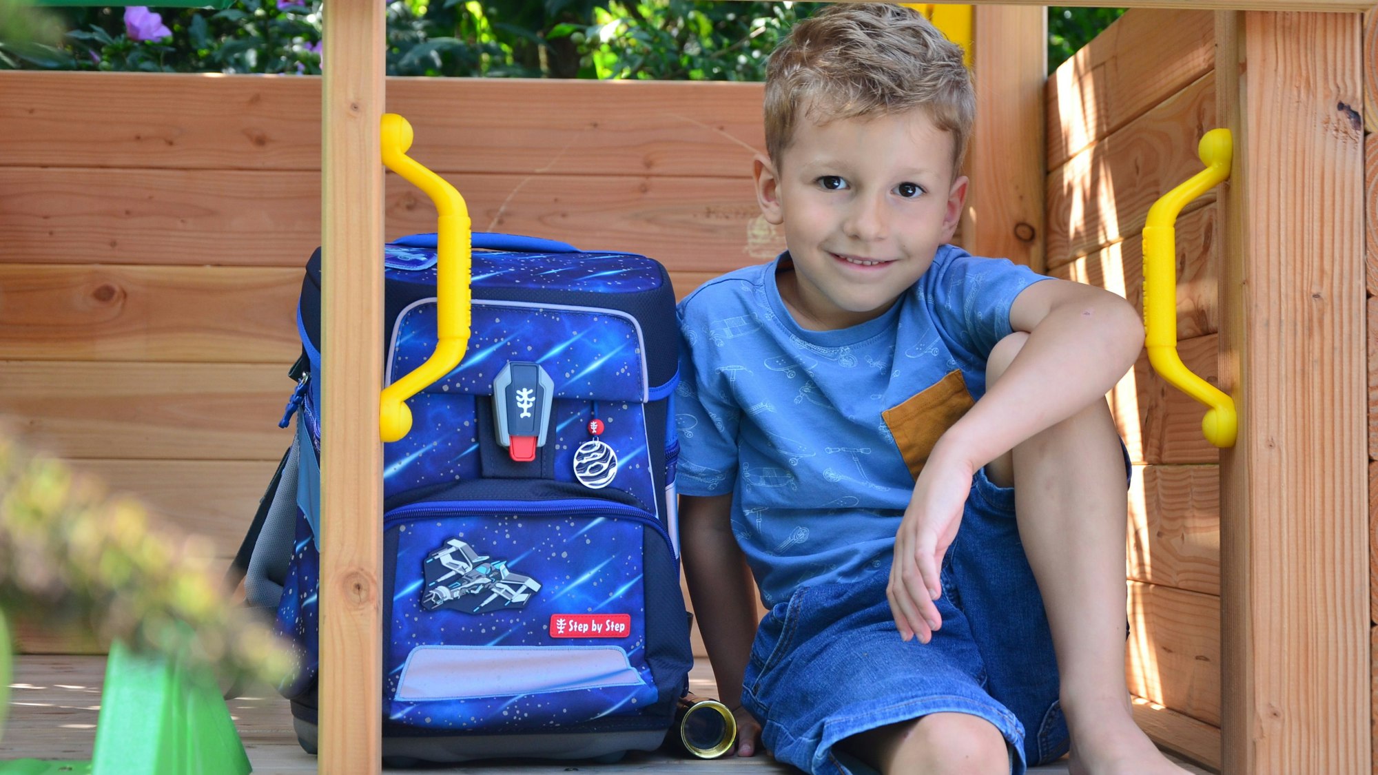Ein Junge sitzt mit einem Schulranzen in einem Baumhaus aus Holz.