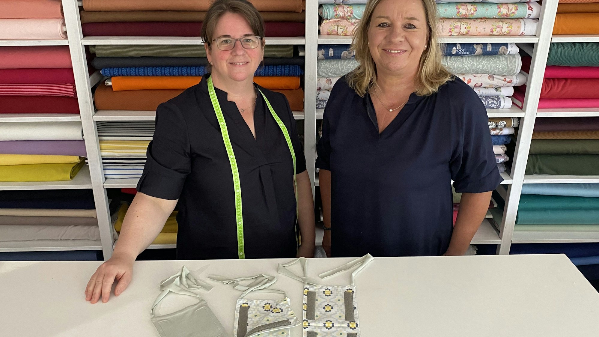 Zwei Frauen stehen vor einem Regal, in dem zahlreiche bunte Stoffballen liegen. Vor ihnen auf dem Tisch liegen mehrere kleine Taschen.