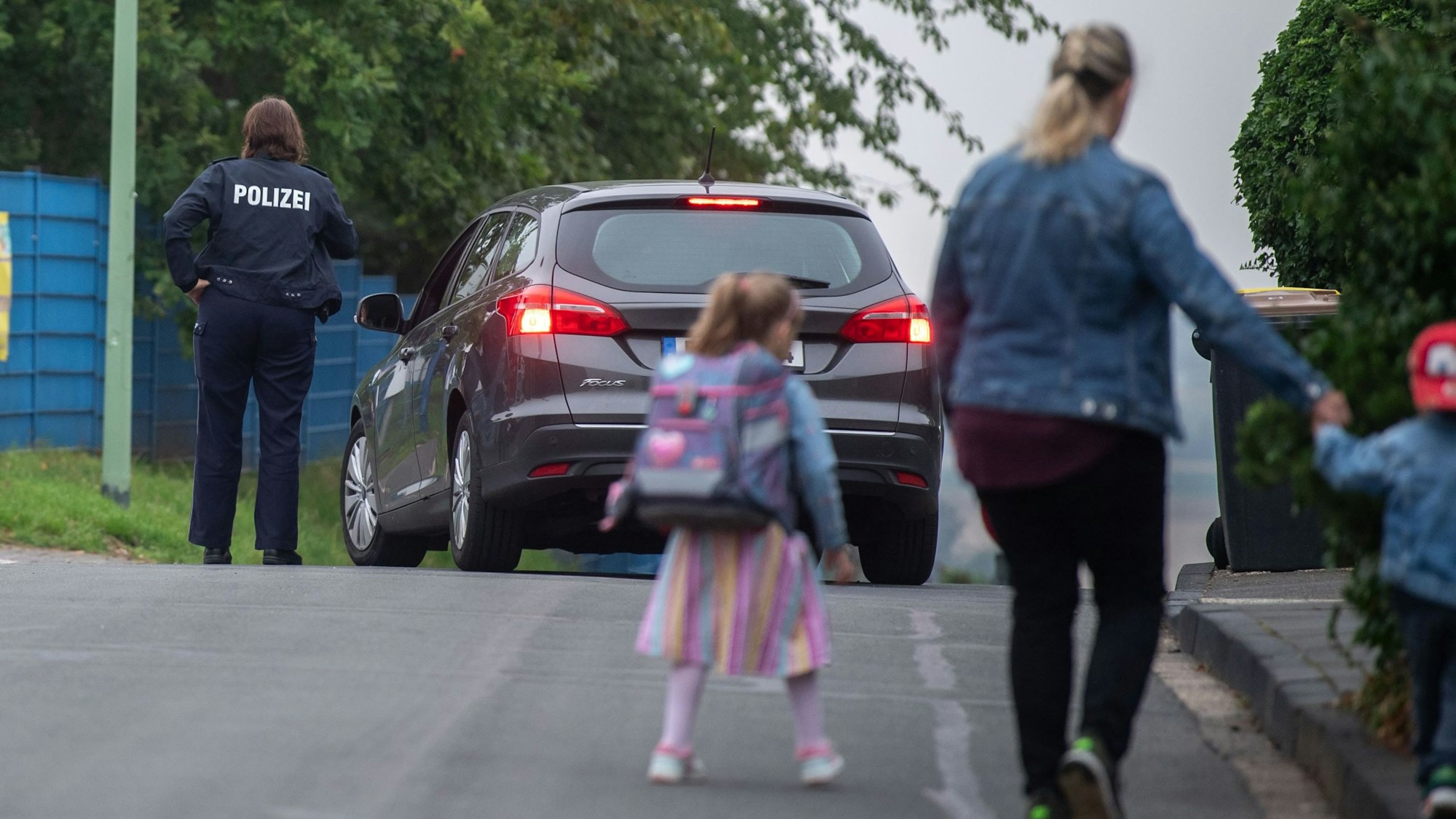 Eine Frau geht mit einem Kind über die Straße. Im Hintergrund sieht man ein Auto und eine Polizistin.