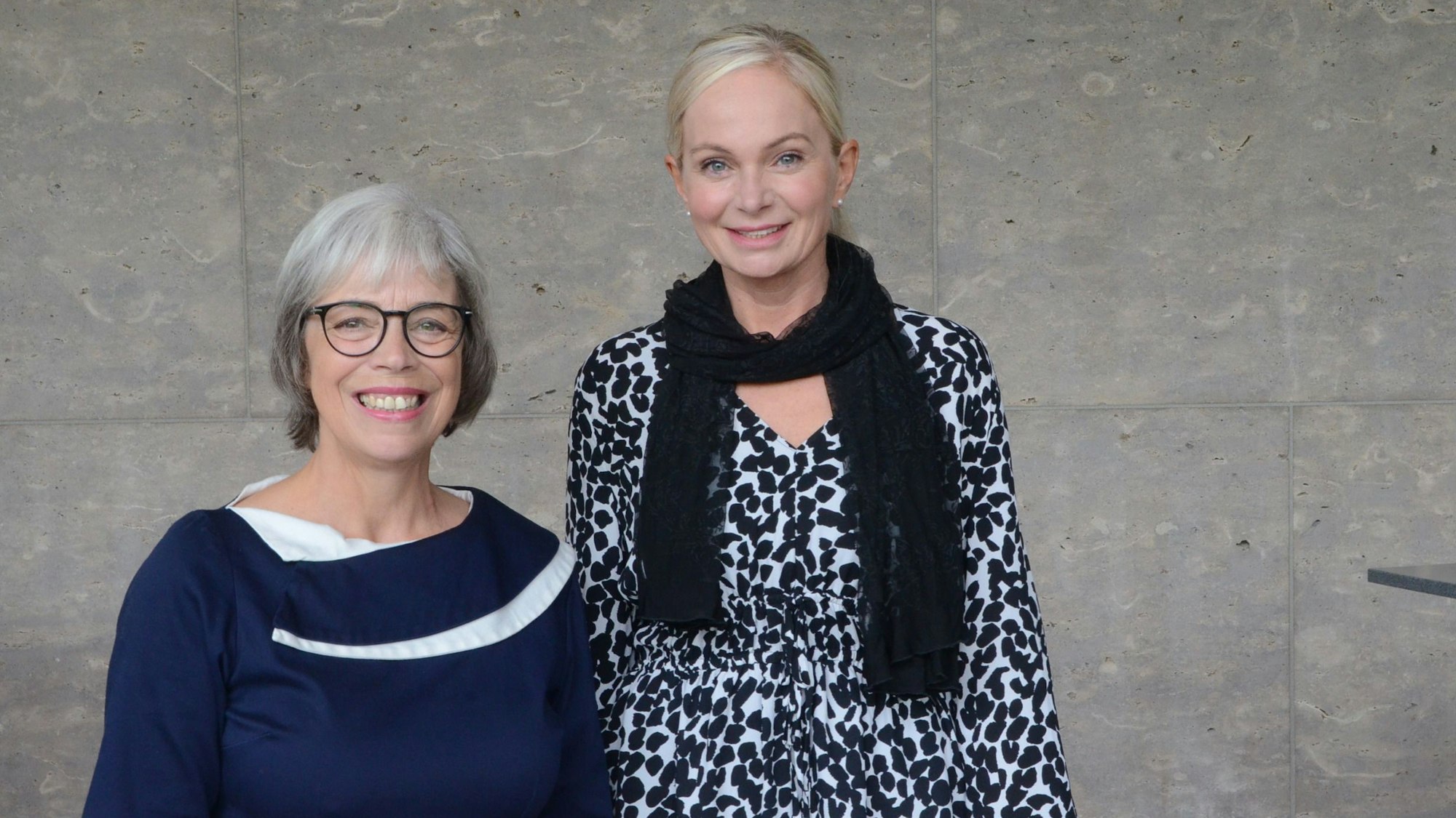 Zwei Frauen vor einer grauen Natursteinwand; die eine hat graues Haar und trägt Brille und eine blaues Kleid, die andere ist deutlich größer, blond und trägt eine schwarz-weiß gemusterte Bluse.