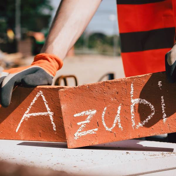 Ein Mann hält in jeder Hand einen Backstein, auf einem steht ein „A“, auf dem anderen der Schriftzug „zubi“.