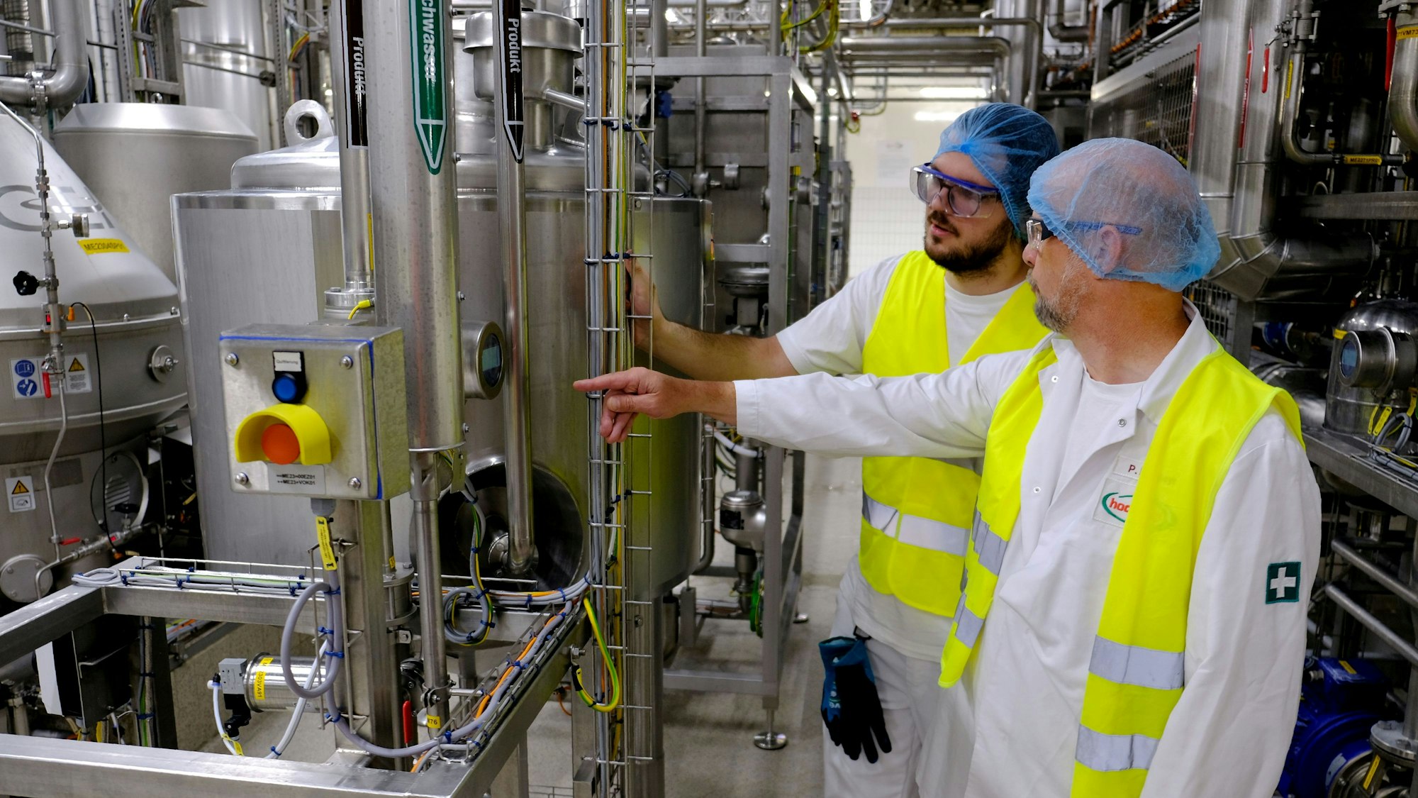 Ausbilder Peter Eiblmayr (r.) und Azubi Nicolas Bertram stehen in weißer Arbeitskleidung mit Haarnetz und gelber Sicherheitsweste in einem Gewirr von Edelstahl-Rohrleitungen einer Molkerei.