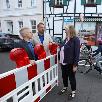 Zwei Männer und eine Frau stehen an einer rot-weißen Sperrbake, im Hintergrund fährt ein Schüler auf dem Fahrrad vorbei.