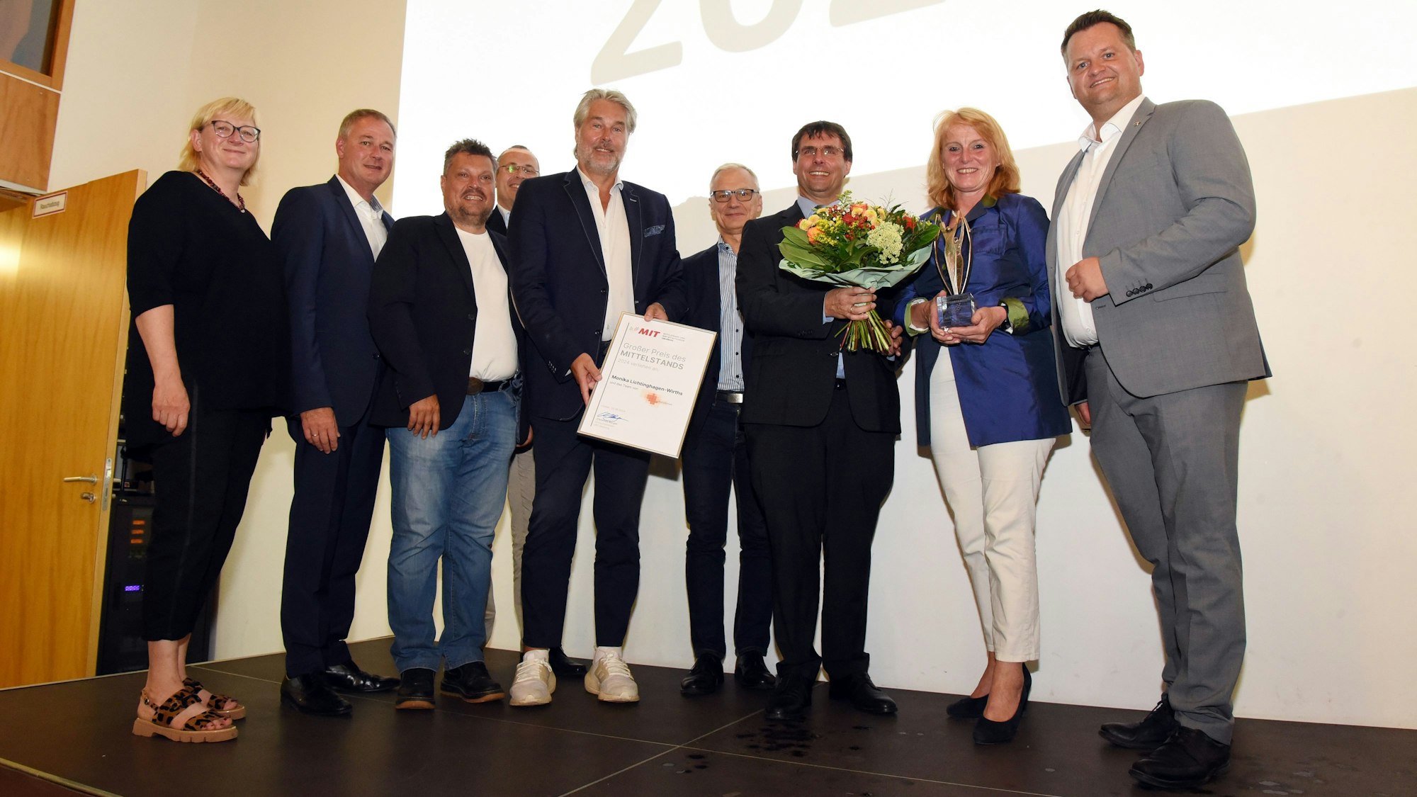 Frauen und Männer stehen in einer Reihe zusammen. Eine Frau hält einen Blumenstrauß und einen Wirtschaftspreis in den Händen.