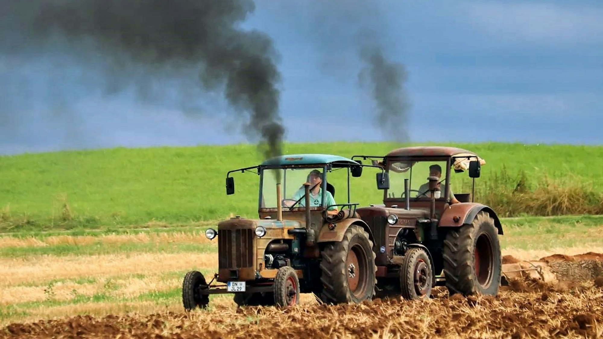 Die beiden Schlepper, aus deren Auspuff dunkler Rauch quillt, ziehen auf dem Acker Baumstämme hinter sich her.
