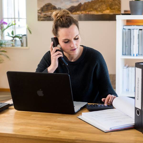 Eine Person sitzt am Schreibtisch und telefoniert. Vor der Person ist ein Laptop zu sehen und im Hintergrund sind Aktenordner in einem Regal.