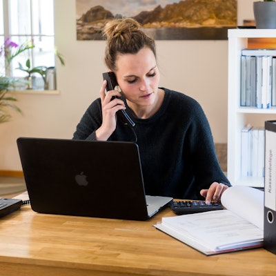 Eine Person sitzt am Schreibtisch und telefoniert. Vor der Person ist ein Laptop zu sehen und im Hintergrund sind Aktenordner in einem Regal.