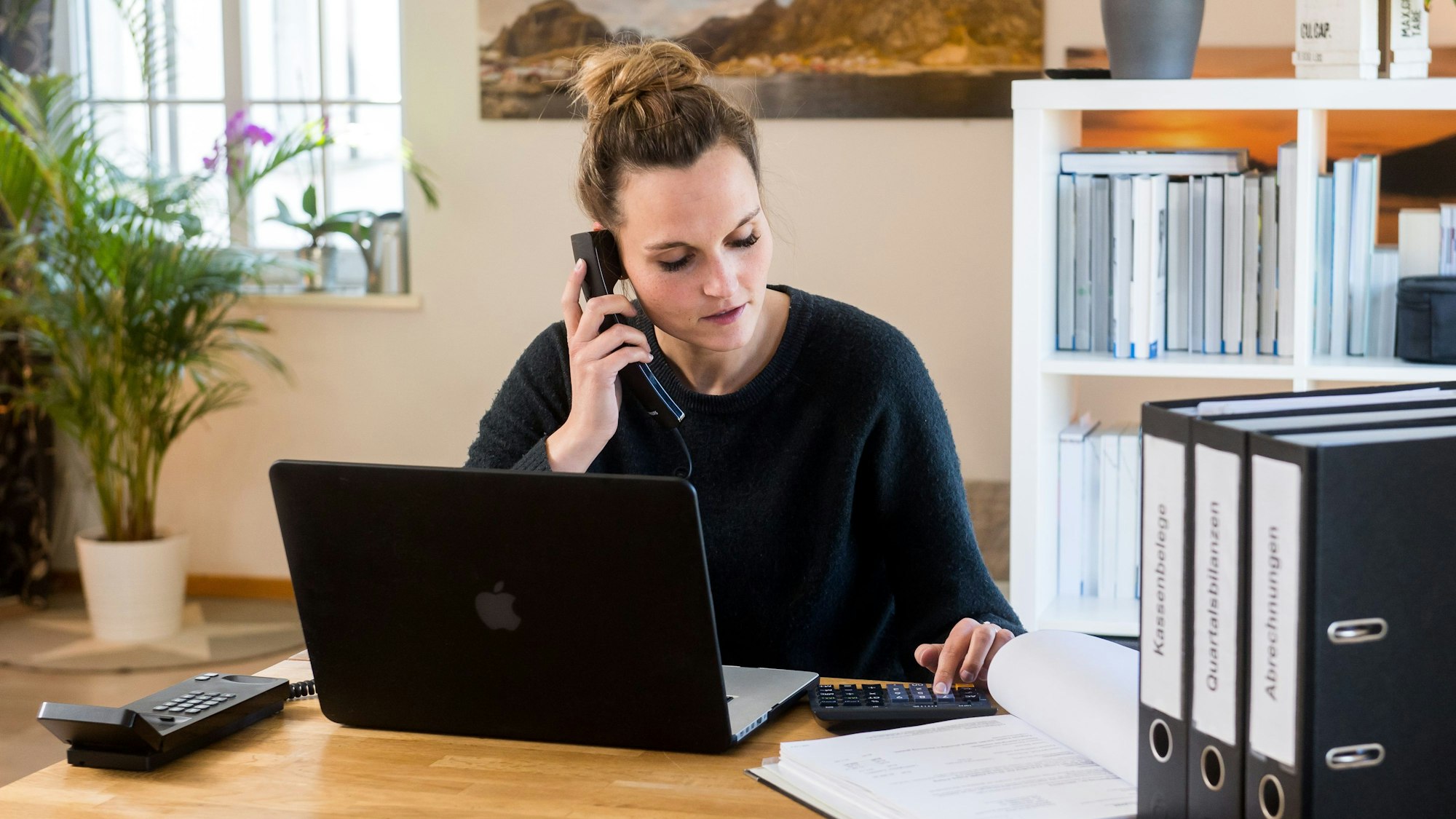 Eine Person sitzt am Schreibtisch und telefoniert. Vor der Person ist ein Laptop zu sehen und im Hintergrund sind Aktenordner in einem Regal.
